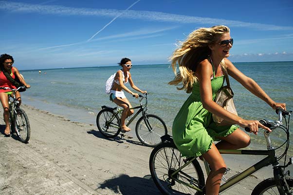 Three young women riding their bikes along the shore.