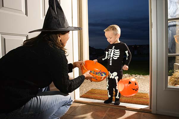 Young boy dressed as a skeleton taking candy at a woman's door for Halloween trick-or-treating.