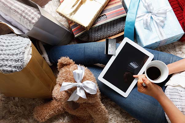 A woman takes a coffee break from online gift shopping surrounded by wrapped presents.