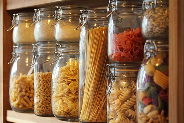 A kitchen pantry shelf with jars of dry beans, pasta and flour.