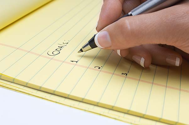 A woman's hand shown with pen writing a list of goals on a yellow legal pad