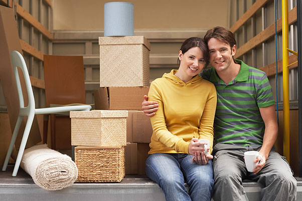 A woman and man sit on the open back of their half loaded moving truck with coffee mugs taking a break from moving