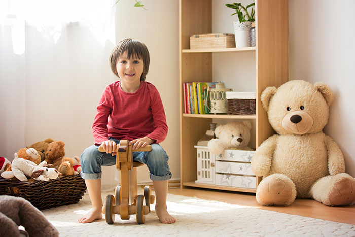A young boy sitting on a toy horse in his playroom surrounded by neat shelves, organized toy bins, books and stuffed animals