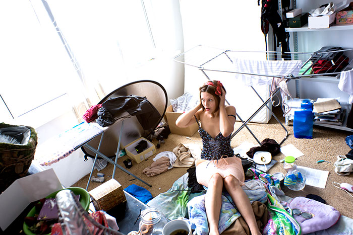 Overwhelmed unorganized woman sitting in her messy room surrounded by clothing and household items.