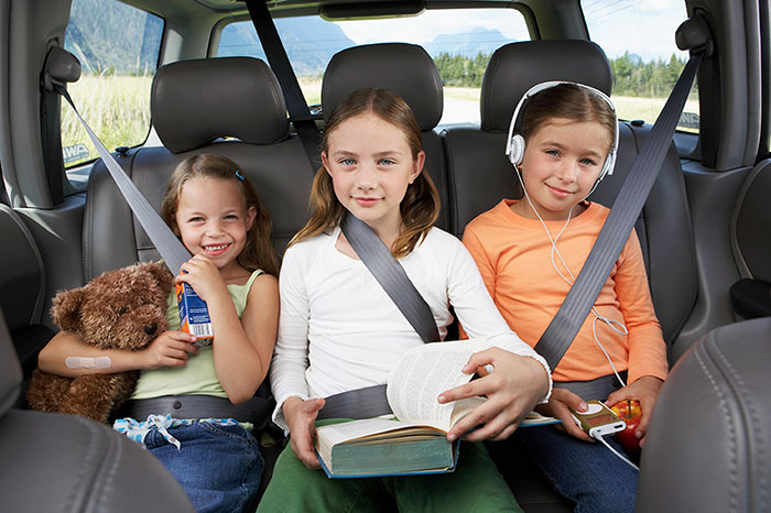 Three young sisters sitting in the back seat on a road trip with the family; reading, listening to music, holding a stuffed animal