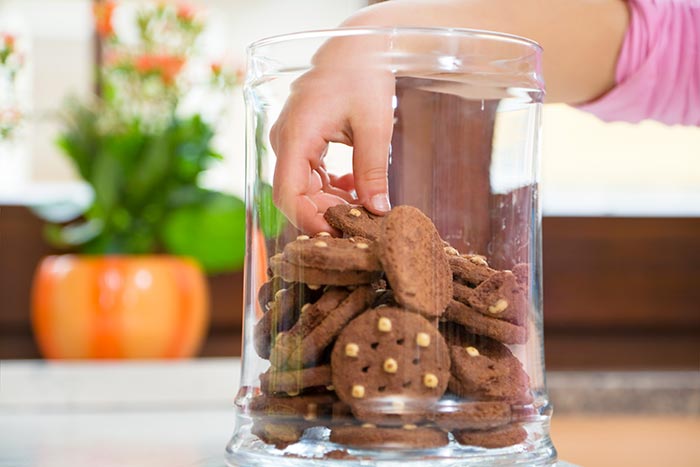 A child reaching hand in a glass cookie jar filled with chocolate macadamia nut cookies sitting on the kitchen counter