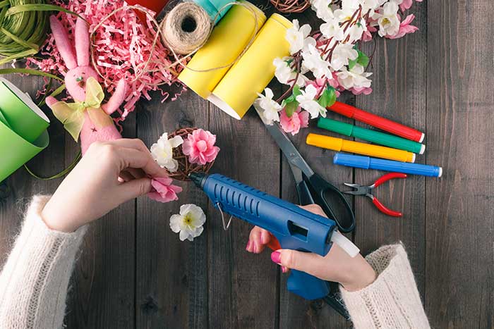 View looking down at craft table, hands hot gluing flowers, with paper, ribbon and scissors strewn on wooden table.