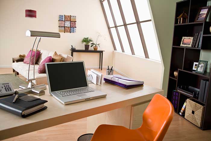 Close up modern office wood desk with bright orange chair, laptop, letter caddy, notebook and bookshelf with sofa and large window in the background