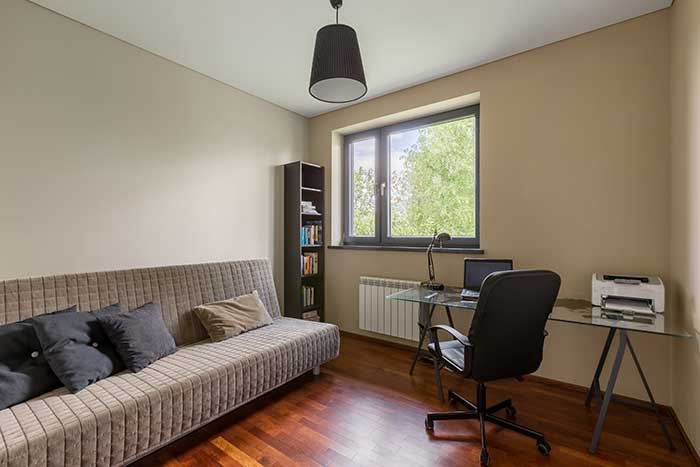 Modern home office with dark wood floors, sofa, desk with laptop and printer, narrow wood bookshelf next to a quaint window
