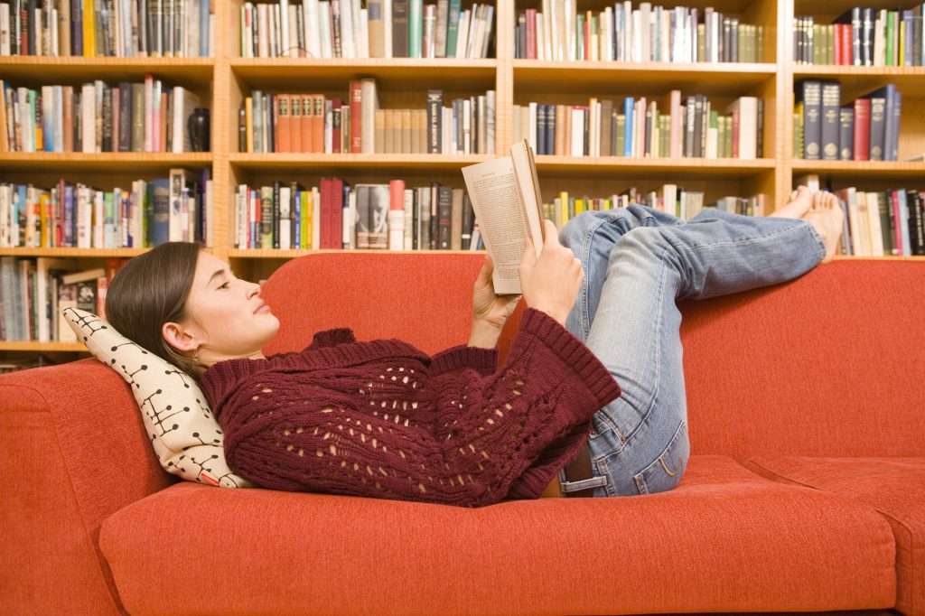 Young woman laying on a couch reading a book in front of a massive bookshelf wall