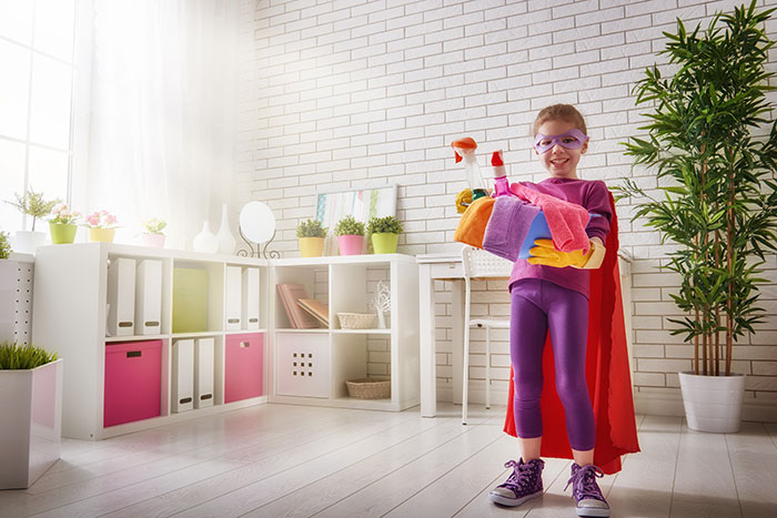 Young girl dressed in purple sneakers and a red cape, holding cleaning supplies ready to tackle cleaning her room