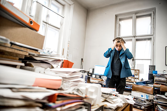 Young man looking frazzled in his cluttered office with papers piled everywhere holding his head