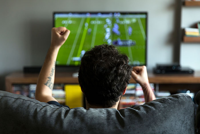 Backside view of man raising his fist to cheer his team while watching a football game on a big screen tv on the sofa in his man cave